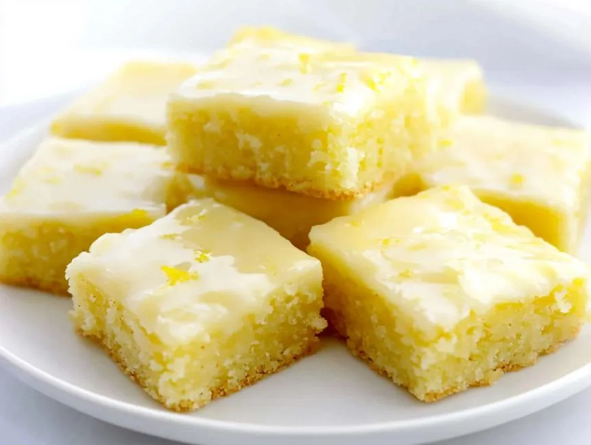 lemon brownies presented as neatly cut squares with a vibrant yellow hue, indicating a moist and tender texture. The base layer appears soft and cake-like with a slightly dense crumb, while the top is coated with a smooth, glossy lemon glaze that has a slightly translucent quality. 