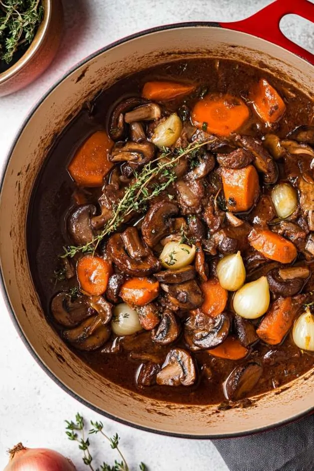 Hearty vegetable bourguignon in a red Dutch oven, featuring tender carrots, mushrooms, pearl onions, and herbs in a rich red wine sauce. Fresh thyme and bay leaves add depth to this savory, slow-cooked dish. A bowl of creamy mashed potatoes sits nearby, ready to be served.